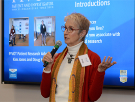 Woman with short grey hair and glasses speaks into a microphone in front of presentation screen