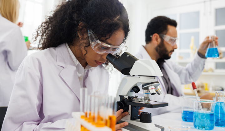 Young, black woman with curly hair in ponytail and safety goggles looks into a microscope