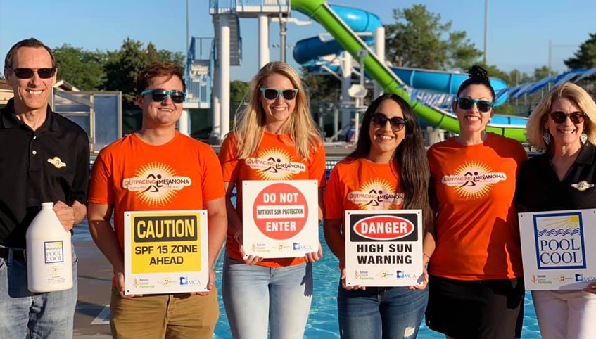 A group of people in sunglasses pose in front of a pool holding sun safety signs