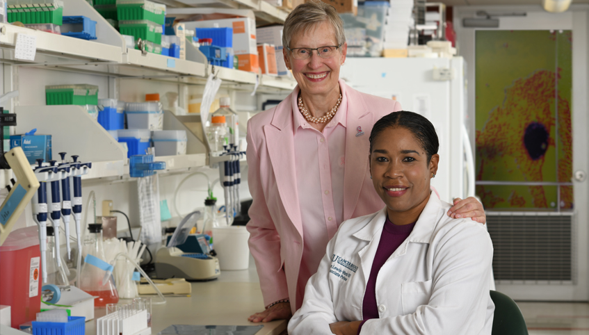 Woman in pink suit stands behind woman researcher in lab coat as she sits at the lab bench