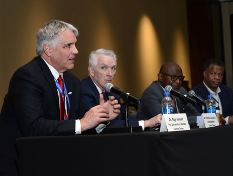 Four men sit at a panel table speaking into microphones