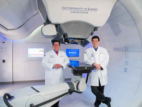 Two doctors stand at proton machine smiling toward the camera