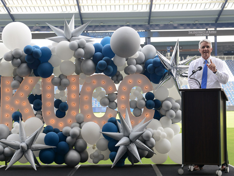 Man in blue tie with grey hair speaks at a lectern in front of light up letters and balloons spelling KUCC