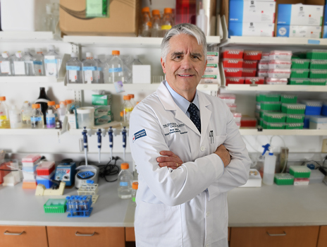Dr. Roy Jensen stands in front of a lab shelf wearing his white lab coat