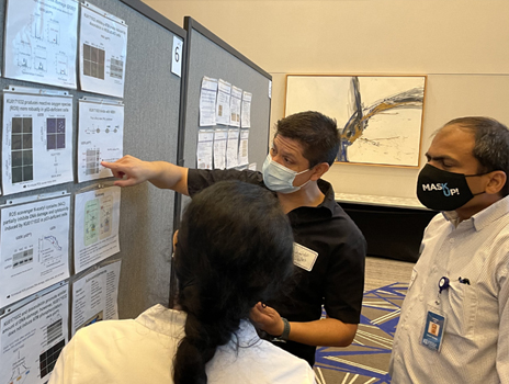Two individuals listen to man in mask pointing to his research poster