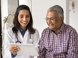 A smiling woman in a white lab coat with a stethoscope around her neck is holding a clipboard, showing it to a gray-haired person wearing glasses and a button down plaid shirt.. They are also smiling 