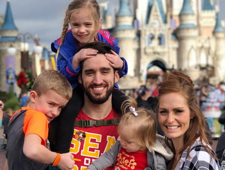A young man with brown hair and beard smiles while surround be his family, one of his daughters on his shoulders smiling while he carries his son. A young woman with light brown hair stands close to him holding their other daughter. Disney's Cinderella's Castle is in the background.