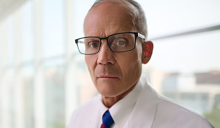 Dr. Joseph McGuirk, Division Director, Hematologic Malignancies and Cellular Therapies at T he University of Kansas Cancer Center is pictured looking straight at the viewer. In the head and shoulders photo of Dr. McGuirk, he has a serious facial expression, is wearing black frame eyeglasses, a shirt and tie and his white coat.