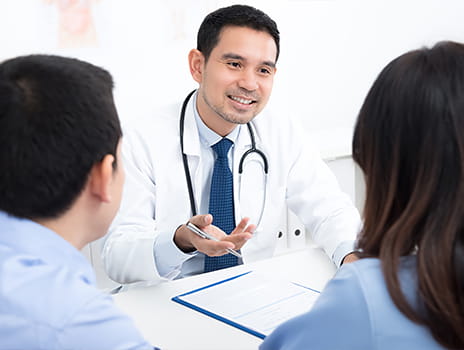 Healthcare professional in a white coat consulting with two patients at a desk, gesturing while discussing a document on a clipboard in a clinical office setting.