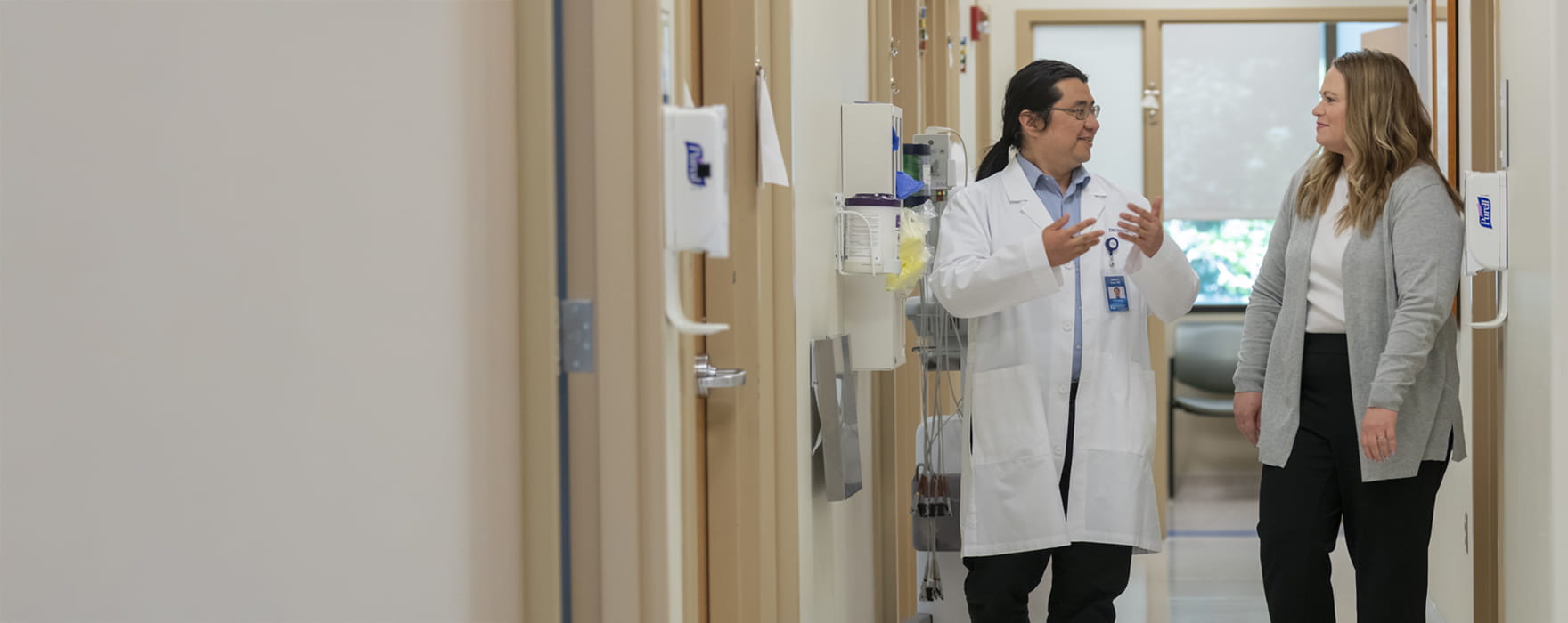 Doctor in white lab coat and woman in grey sweater talk while walking down a clinical hallway