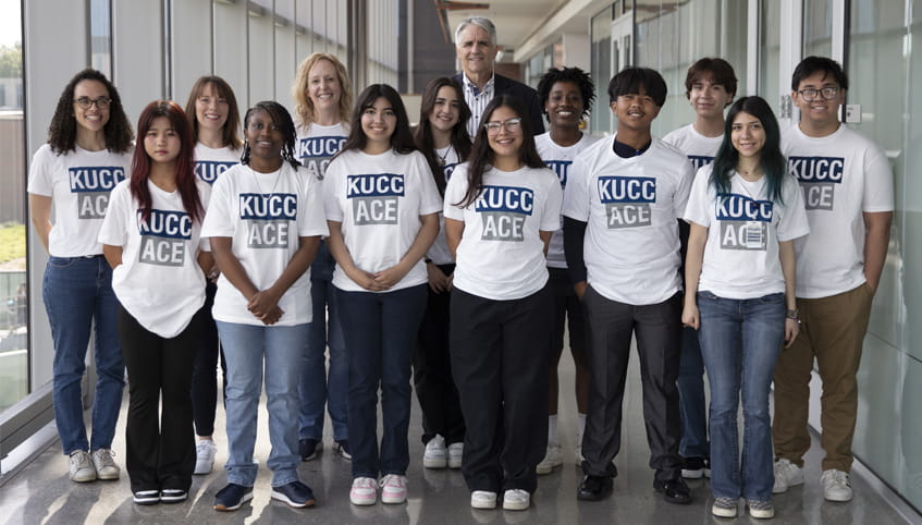 Group of young people stand together smiling toward the camera, they are wearing white shirts that say "KUCC ACE" on the front.