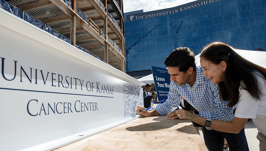 Two people sign a white steel beam in front of a modern building