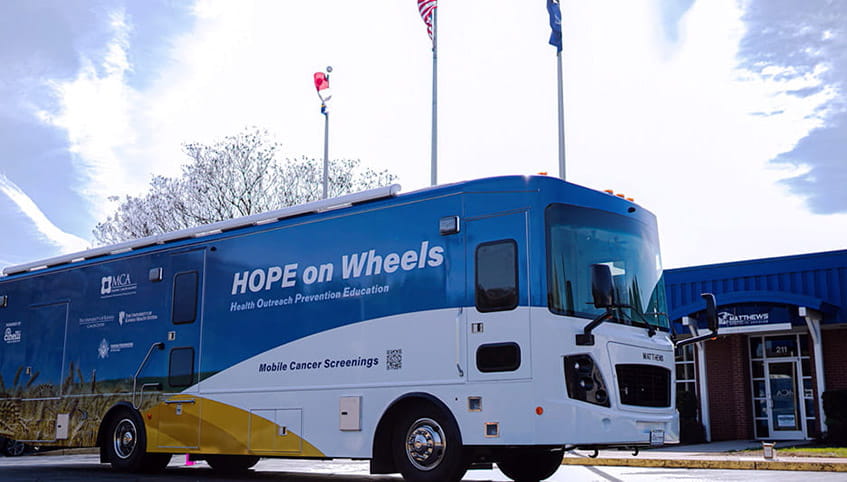 The Masonic Cancer Alliance mobile screening unit, Hope on Wheels, sits in a parking lot at the front door of a one-story brick building. The sky is bright blue with a large white cloud. 