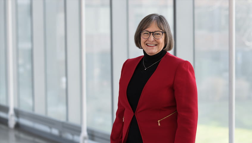 Dr. Maliski stands in a red coat in front of a wall of windows smiling at the camera