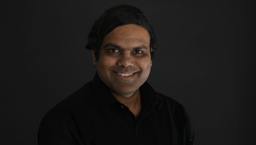 Man with dark hair smiles at the camera in front of a dark grey background.