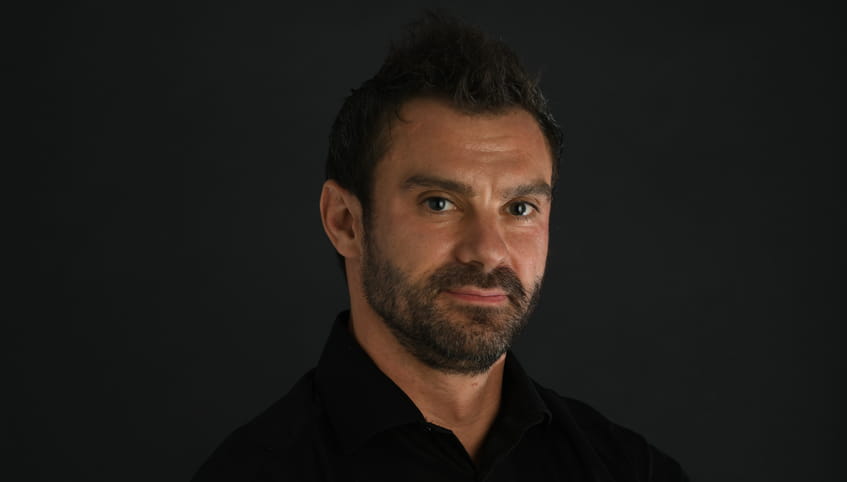 Man with dark hair and beard smiles at camera with a grey background.