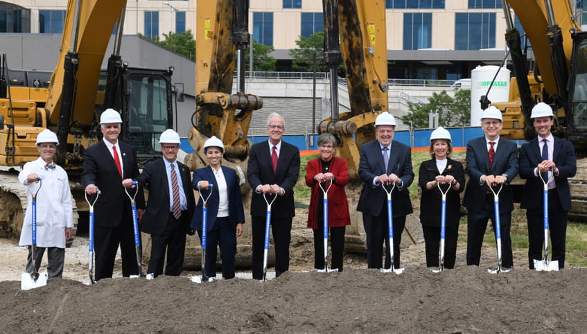 Group of individuals - several in suits and one in a lab coat - standing in front of large, yellow construction equipment hold shovels while wearing hard hats. 