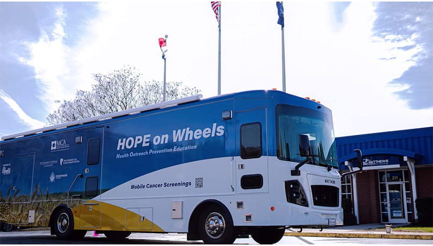 A large RV is parked outside a community clinic with a bright blue sky and white cloud above. The side of the RV has the phrase "HOPE on Wheels".