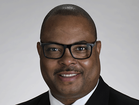 Babalola Faseru poses for a professional headshot. They wear a black suit, white shirt, yellow tie, and glasses.