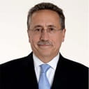 Headshot of a person wearing black coat, white shirt, blue tie, glasses and a mustache.