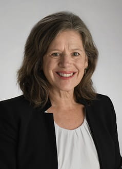a professional headshot of a person smiling while wearing a black blazer over a white shirt and in front of a gray background