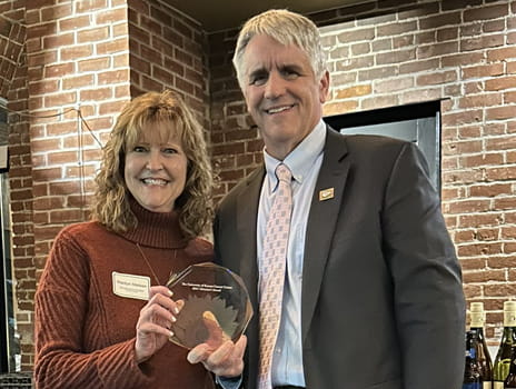 Two individuals holding an award at an indoor event smiling before a brick wall background.