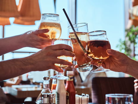 Four people clinking glasses filled with various drinks at a sunlit restaurant table, celebrating or toasting together.