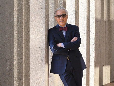 Man wearing glasses and a suit leans against a wall smiling toward the camera