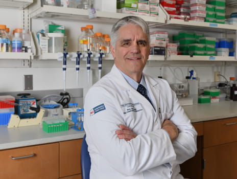 Main in white coat stands with arms crossed smiling at the camera