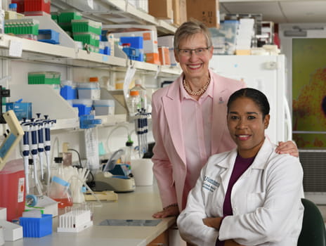 A woman in pink stands behind a sitting woman in a lab coat in front of a laboratory bench