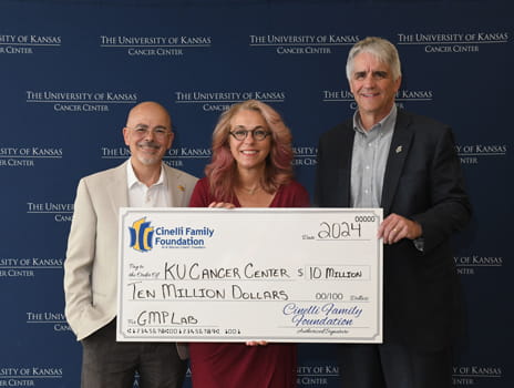 Three people stand holding a large check made to KU Cancer Center for $10 million.