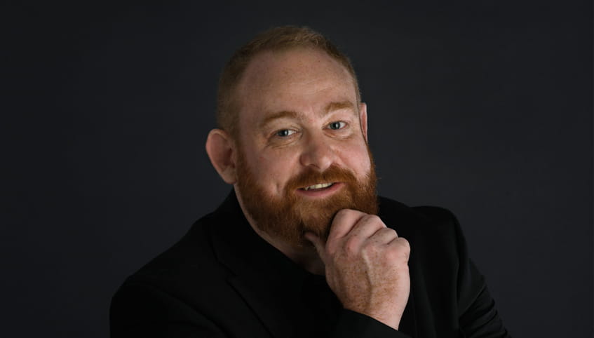 A person with red hair and a beard smiles while resting his chin on his hand.