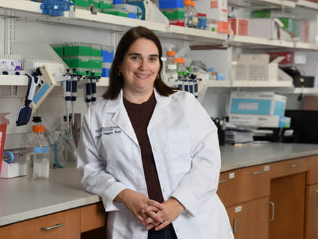 Person stands in front of a science lab bench wearing a white lab coat.