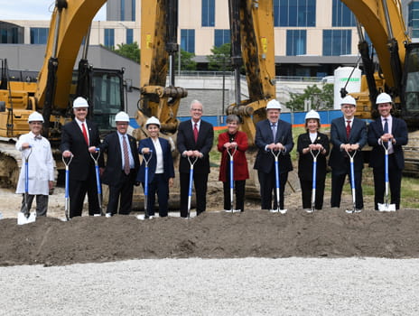 A group of people stand in front of construction equipment holding shovels. Individuals are in business dress and lab coats and are wearing hard hats. 