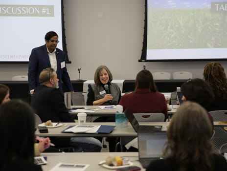 Woman speaks into microphone while seated in a group of people who listen attentively in a classroom. 