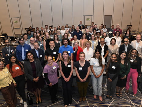 A large group of people wearing nametags stands together for a group photo inside a convention center hall. 