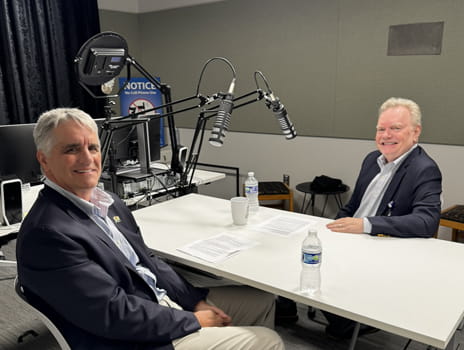 Two men in sport coats sit in a podcast studio in front of microphones. 