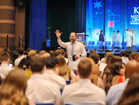 A man stands in a large crowd waving with a blue stage lit up behind him. 