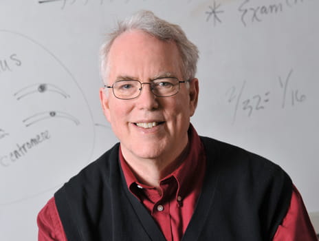 A man smiling with glasses and grey hair wearing a red shirt and black vest in front of a whiteboard with equations written on it. 