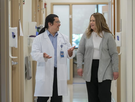 A doctor in a white lab coat walks and talks with a female patient in a medical facility hallway.