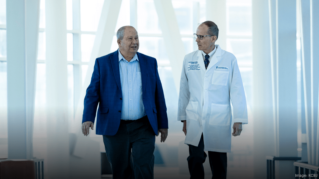 David Frantze, a patient at The University of Kansas Cancer Center, is wearing a blue suit and white, open collar, button down shirt while walking and speaking with Joseph McGuirk, DO, Hematologic Malignancies and Cellular Therapeutics, who is wearing his white lab coat.