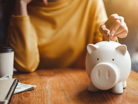 A porcelain, white piggy bank sits on wooden table next to notebooks and a cup of coffee, while a women in the background wearing an orange shirt places a coin in it