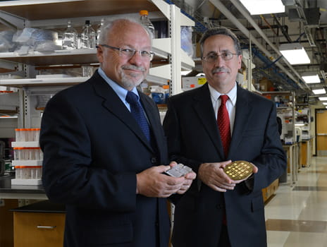 Two men wearing suits and glasses stand in a scientific lab holding lab equipment