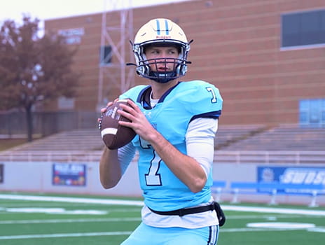 American football player in a light blue number 7 jersey holding a football mid‑throw on an outdoor field with bleachers and a brick building in the background.