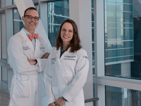 Two people dressed in white coats stand in a modern hallway with glass windows while smiling.