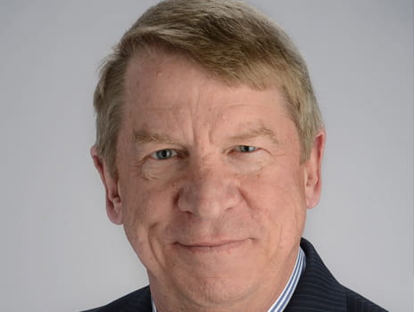 Close-up portrait of an individual with a confident smile, wearing a dark blazer. The background is plain and light-colored.