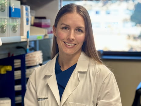 A professional wearing a white lab coat, smiling in a laboratory setting, with shelves stocked with supplies in the background.