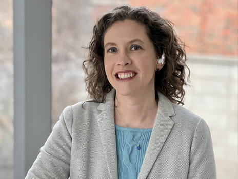 Person with brown, curly, shoulder length hair smiles at the camera. They are wearing a blue shirt with grey sport coat and stand in front of a large window. 