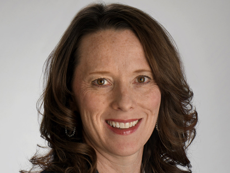 Close-up portrait of a smiling person with long, curly brown hair, wearing earrings, against a light gray background.