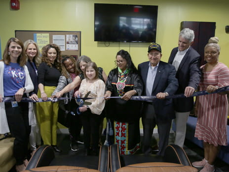 A group of people holds a blue, while a girl with large scissors cuts it